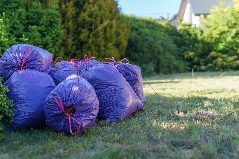 A row of several large purple garden waste bags, tied securely with red ribbons, are placed on a patch of grass in a backyard for garden maintenance or landscaping work. The bags are full of garden de